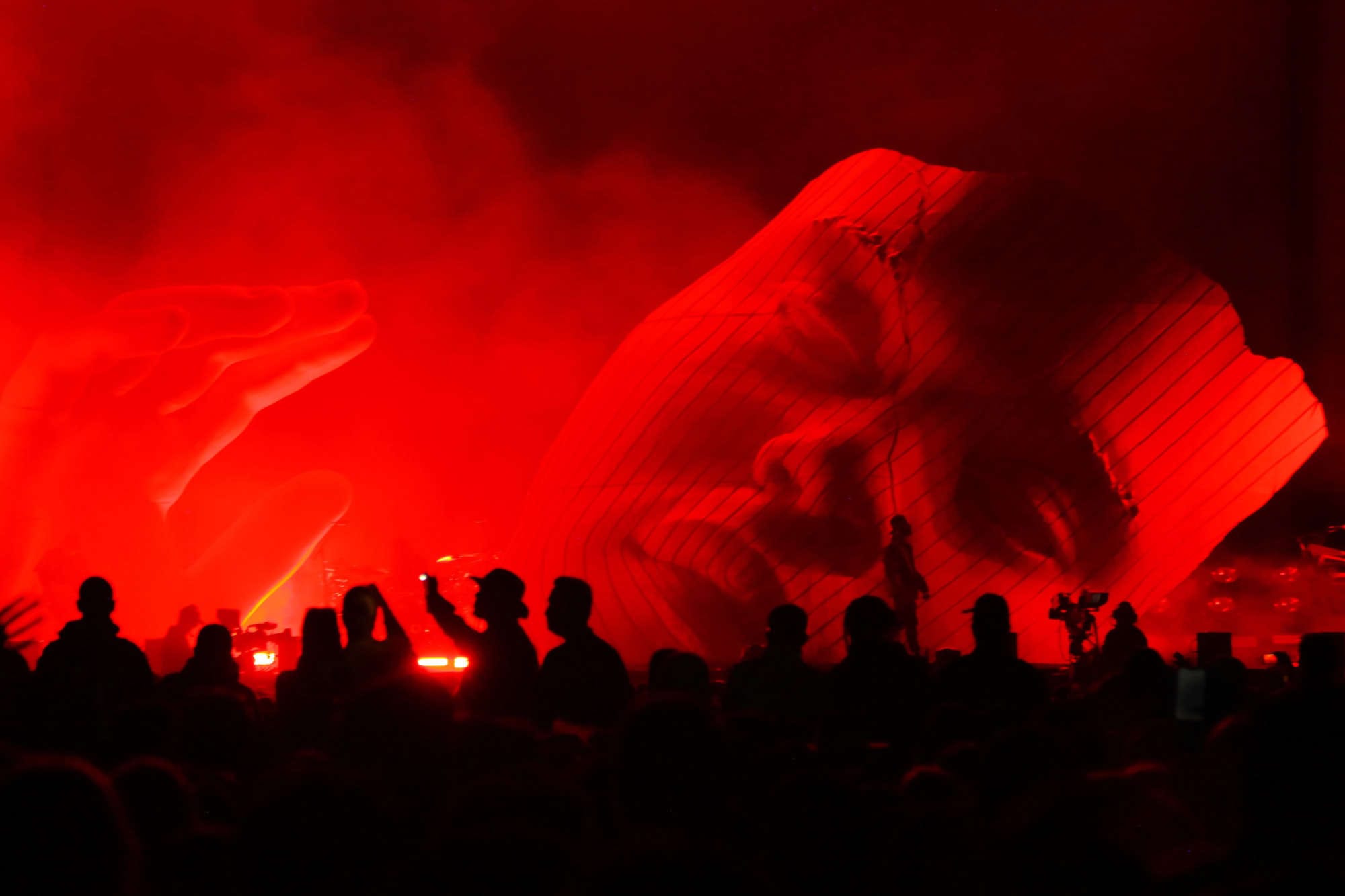 Es Devlin stage design with a monumental red sculpted face and silhouetted audience during a live performance, illustrating her immersive, human-scale approach to light and architecture.