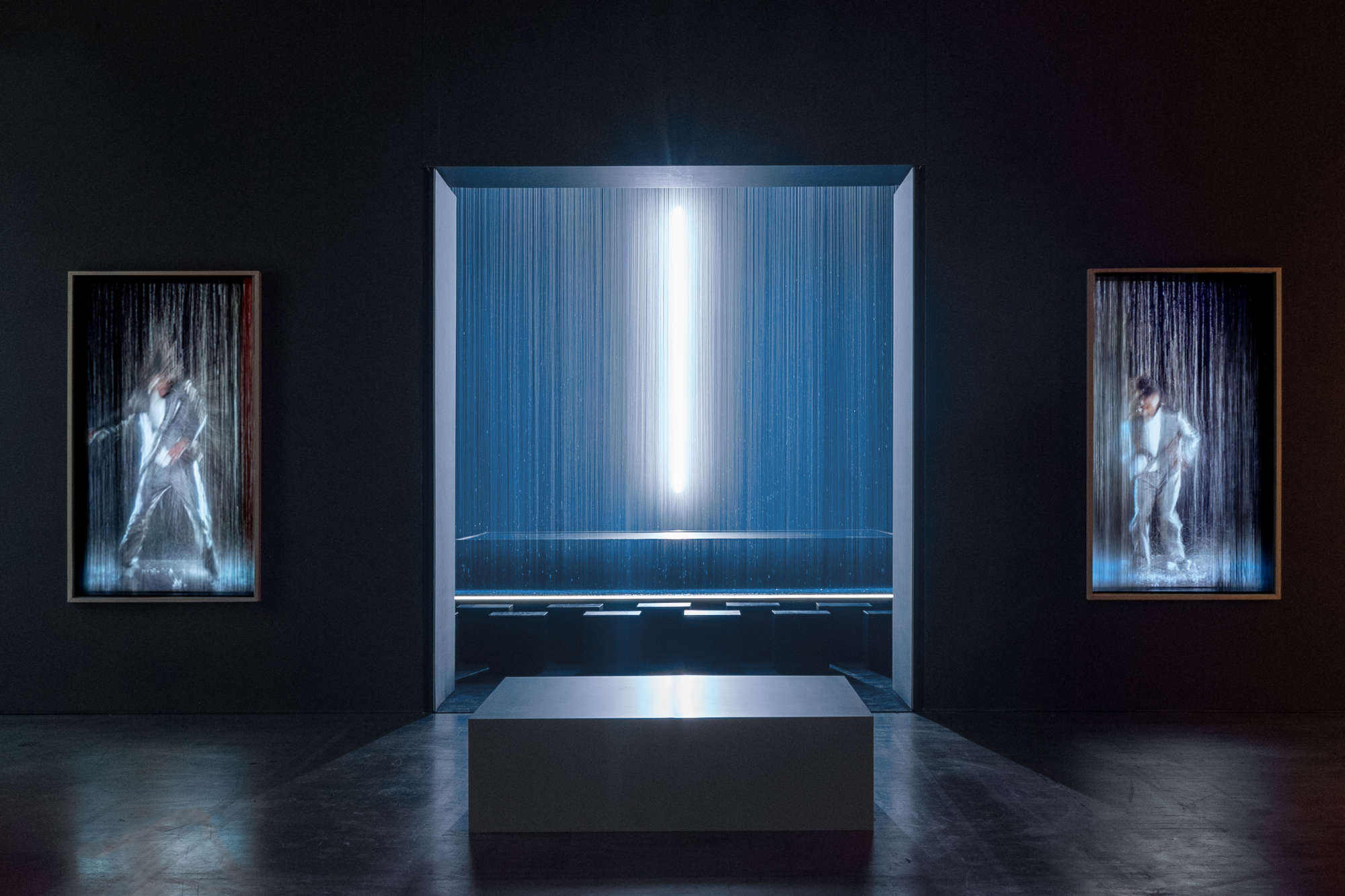 Es Devlin light installation featuring a glowing vertical beam suspended within cascading threads of light, flanked by two illuminated portraits in motion — an immersive study of reflection, rhythm, and presence.